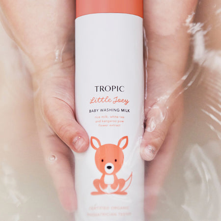 The image features a bottle of Tropic Little Joey Baby Washing Milk being held gently in water. The packaging is white with a playful orange kangaroo illustration, emphasizing its suitability for babies. Key ingredients, including rice milk, white tea, and kangaroo paw flower extract, are highlighted on the label. The product is Certified Organic and Pediatrician Tested, ensuring it is safe and gentle for delicate baby skin.